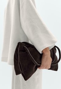Sac à main en suède marron avec un design plié, présentant des bords doux et une seule anse, tenu par une personne portant un haut en lin blanc ample.