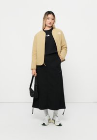 Blouson bomber beige clair sur un sweat-shirt noir, associé à une longue jupe noire. Les accessoires comprennent un sac noir et des chaussettes grises avec des chaussures blanches.
