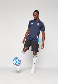 Camisa deportiva azul marino con acentos rojos y azules, pantalones cortos negros, calcetines blancos con tres rayas negras y un balón de fútbol azul con estampado.