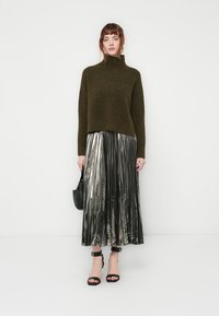 Maglione a collo alto in verde oliva abbinato a una gonna a pieghe metalliche argentate, con tacchi a listini neri e una piccola borsa nera.