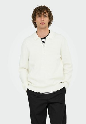 Suéter blanco de cremallera con canales y cuello, usado sobre una camisa de rayas blancas y negras, combinado con pantalones negros.
