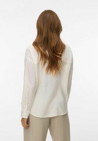 Femme aux longs cheveux bruns ondulés portant une chemise blanche ample à manches longues et un pantalon beige, debout de dos sur un fond blanc.
