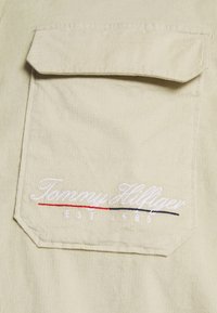 Beżowa tkanina z sztruksu z dużą kieszenią na piersi oraz haftowanym napisem "Tommy Hilfiger" z czerwonymi i niebieskimi akcentami. Zawiera również napis "EST. 1985".