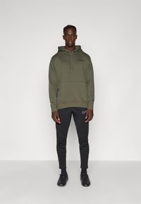 Felpa verde oliva con cappuccio e lacci, abbinata a pantaloni tuta neri con logo Nike bianco, e scarpe sportive nere.