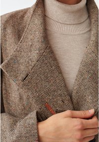 Blazer de tweed marrón con un tejido texturizado y motas multicolores. Presenta un cuello con muesca y un cierre frontal abotonado, superpuesto a un ligero cuello de tortuga.