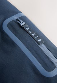 Marineblå stofpose med en glat tekstur, der har en kontrasterende blå lynlås og en etiket med "BAKER" i hvide bogstaver.