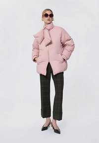 Γυναίκα φορώντας μια υπέρμετρα ροζ φουσκωτή ζακέτα με δεμένο κασκόλ, σκούρες καρό παντελόνες, μαύρες ψηλές γόβες και μεγάλα γυαλιά ηλίου, στέκεται με τα χέρια στις τσέπες.