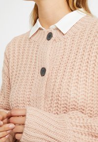 Ljusrosa chunky stickad cardigan med två stora mörka knappar, bärs över en vit skjorta med krage, händer delvis synliga framifrån.