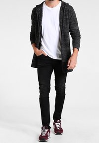 Cardigan lungo grigio scuro, texture, con cappuccio, indossato sopra una maglietta bianca, abbinato a jeans neri slim-fit e sneakers atletiche bordeaux.