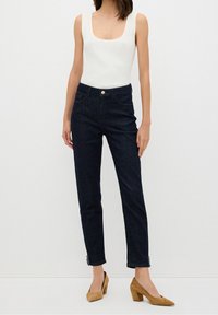 Femme portant un haut blanc sans manches, un jean bleu foncé taille haute avec des ourlets décoratifs, et des chaussures à talons blocs couleur beige, debout devant un fond blanc.