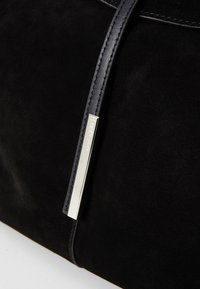 Zwarte suède handtas met een gladde afwerking, voorzien van een strakke zilveren logo-accent en lederen handvat met stiksels. Minimalistisch ontwerp, geen zichtbare patronen.