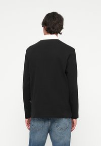 Melns kreklis ar garām piedurknēm un baltu apkakli, izgatavots no gludas auduma. Iezīmējas ar brīvu piegriezumu un vienkāršu dizainu.