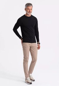 Musta pitkähihaine paita, beige kapea housut ja vaaleat lenkkarit. MAlli seisoo kädet lanteilla, esittäen asun istuvuutta ja muotoilua.