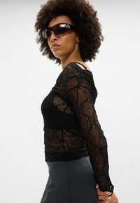 Čierny čipkový top s dlhými rukávmi a zložitými kvetinovými vzormi, doplnený o korálkové akcenty, spárovaný s čiernou koženou sukňou a slnečnými okuliarmi.