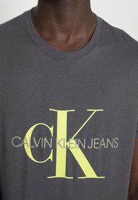 Šedé bavlněné tričko s žlutým a šedým grafickým designem a textem "CALVIN KLEIN JEANS", kulatým výstřihem a krátkými rukávy.