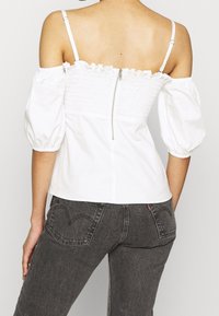 Donna che indossa un top bianco off-shoulder con maniche a sbuffo e chiusura lampo sul retro, abbinato a jeans in denim neri a vita alta.