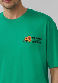 Groene katoenen T-shirt met een grafiek van gesneden papaja en de tekst "PAPAYA smaak SURFWAX" op het linker borstgebied.