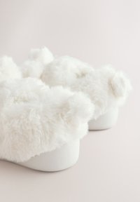 Hvide bløde hjemmesko med en blød, plyset tekstur, rund form og hvide gummisåler; har små pompon-detaljer på toppen.