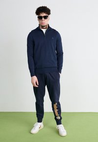 Modra zip-up dukserica, usklađene trenirke s "Timberland" logom, bijele tenisice i sunčane naočale. Tkanina je glatka, s rebrastim manžetama i podstavom.