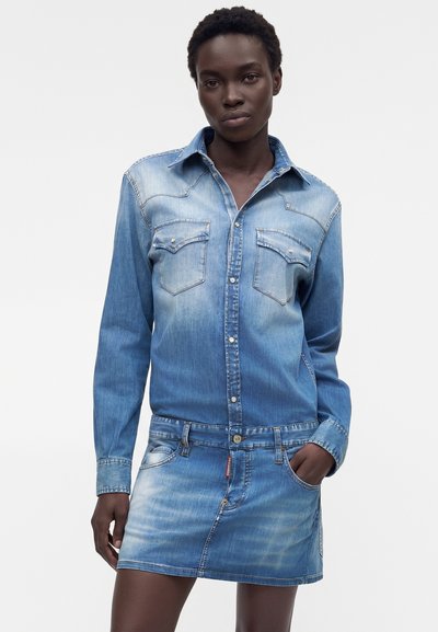 Γυναίκα που φοράει ένα ξεθωριασμένο μπλε τζιν πουκάμισο-φόρεμα με μακριά μανίκια, κουμπωτό μπροστινό μέρος, τσέπες στο στήθος και μια κοντή φούστα με μπροστινές τσέπες.