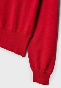 Červený pletený sveter s rebrovanými lemami na rukávoch a spodnom okraji. Mäkká textúra s hladkým povrchom, ktorý vykazuje mierne variácie v priadzi.