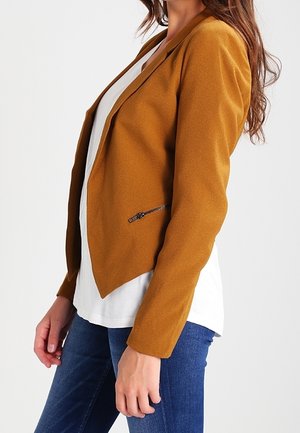 Femme portant un blazer couleur moutarde sur une chemise blanche et un jean bleu, debout devant un fond blanc uni.