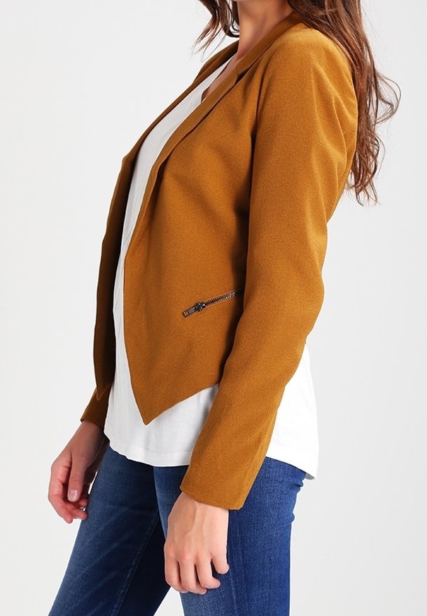 Femme portant un blazer couleur moutarde sur une chemise blanche et un jean bleu, debout devant un fond blanc uni.