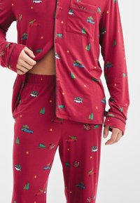 Rood pyjamastel met illustraties van auto's, fietsen en bomen. De stof is zacht met een gladde textuur en een overhemdontwerp met knopen.