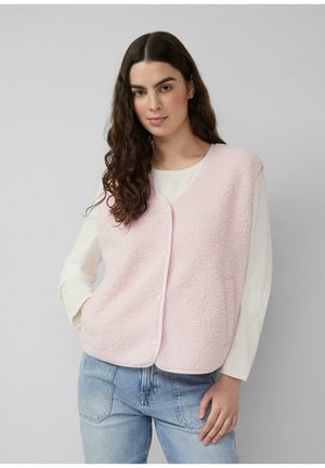 Kobieta o długich ciemnych włosach nosi jasnoróżową kamizelkę z polaru zapinaną na guziki, pod którą ma kremową koszulę z długim rękawem oraz jasnoniebieskie dżinsy, stoi na tle szarego tła.