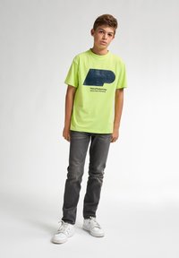 Camiseta verde neón brillante con un logotipo gráfico y texto en azul oscuro. Combinada con jeans gris oscuro y zapatillas blancas. Diseño simple y casual.