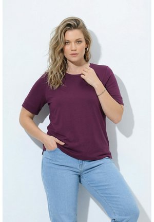 Frau mit langen, welligen blonden Haaren, die ein lila kurzärmeliges Shirt und hellblaue Jeans trägt, steht vor einem weißen Hintergrund.