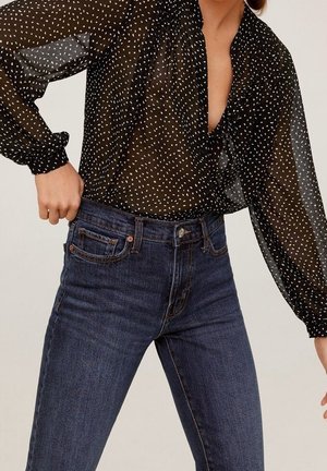 Femme portant un chemisier noir transparent à pois blancs, rentré dans un jean bleu foncé taille haute, ajustant sa ceinture de la main gauche.