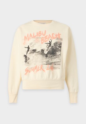 Osoba w okularach przeciwsłonecznych, żółtej bluzie „Malibu Beach”, luźnych ciemnych spodniach i spiczastych butach stoi z jedną ręką w kieszeni.