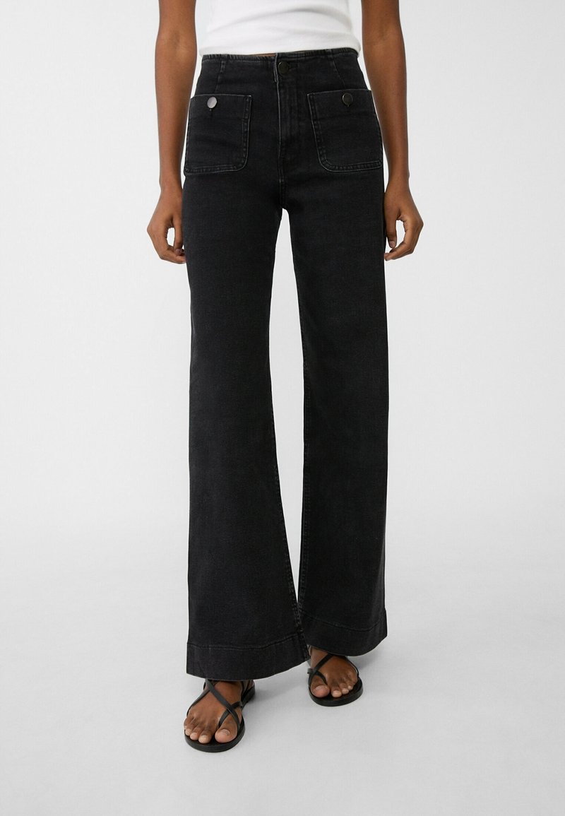 Personne portant un jean noir taille haute à jambes larges avec des poches plaquées à l'avant et des sandales noires à brides, montrée de la taille aux pieds.