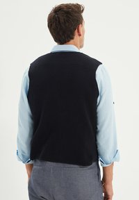 Marinblå stickad väst med en texturerad yta, bärs över en ljusblå skjorta. Vesten har en klassisk design och en avslappnad passform.