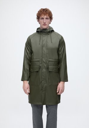 Άνδρας με σγουρά μαλλιά που φοράει πράσινο, αδιάβροχο παλτό μέχρι τα γόνατα με μπροστινές τσέπες και κουκούλα, ο οποίος στέκεται μπροστά σε απλό λευκό φόντο.