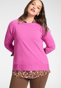 Różowy sweter z dzianiny na brązowej koszuli w panterkę, z okrągłym dekoltem i długimi rękawami. Dopełnieniem stylizacji są brązowe dopasowane spodnie.