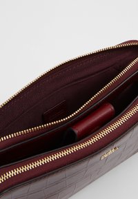 Burgundin värinen nahkalaukku, jossa on tikattu kuvio, kultaiset vetoketjut ja sisäiset taskut, vuorattu tekstuurilla.