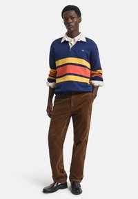Blue rugby shirt with horizontal red, yellow, and orange stripes, paired with brown corduroy pants and dark shoes, featuring a collared design.