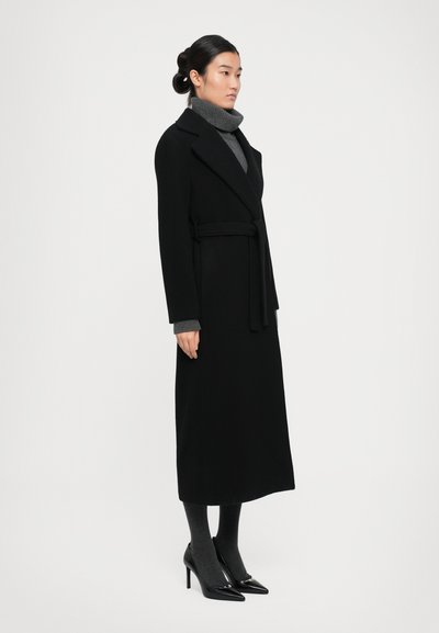 Woman standing in black belted long coat, grey turtleneck sweater, grey tights, and black high-heeled shoes against white background.