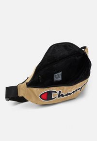 Sac banane en tissu beige avec un grand logo noir et rouge, comportant un compartiment principal zippé et des poches d'organisation intérieures noires.