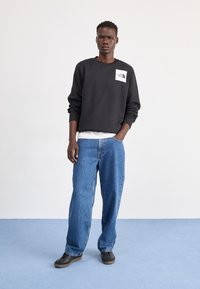 Svart hettegenser med en hvit logo på brystet, kombinert med løse blå denimjeans og svarte sko. Enkelt design, avslappet passform.