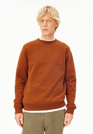 Junger Erwachsener mit blondem Haar, trägt einen rostfarbenen Pullover über einem weißen Hemd und beige Cordhosen vor einem schlichten Hintergrund.