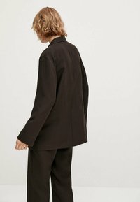 Blazer marron foncé à coupe décontractée, col cranté et manches longues. Tissu texturé, coutures minimales, couture arrière visible.