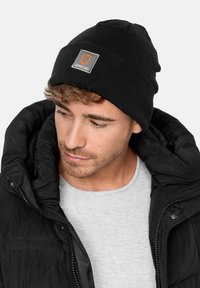 Black knit beanie with a gray patch labeled "JEANSCODE." Worn with a gray sweater and a black puffer jacket, showcasing a textured surface.