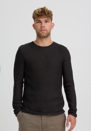 Jeune homme aux cheveux blonds courts et bouclés, portant un t-shirt noir ajusté à manches longues et un pantalon beige, debout devant un fond clair uni.