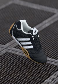 Melni Adidas sporta apavi ar baltām strīpām un gumijas zoli uz metāla restēm, skatīti no augšas leņķī.