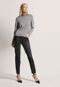 Frau, die einen grauen Rollkragenpullover, schwarze Lederhosen und schwarze High Heels trägt, steht vor einem schlichten hellen Hintergrund.