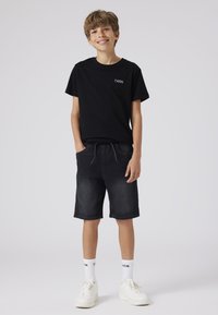 Juoda marškinėlių su logotipu, tamsūs džinsiniai šortai su raišteliu, balti sportbačiai ir baltos kojinės su juodu tekstu, pateikti ant paprasto fono.