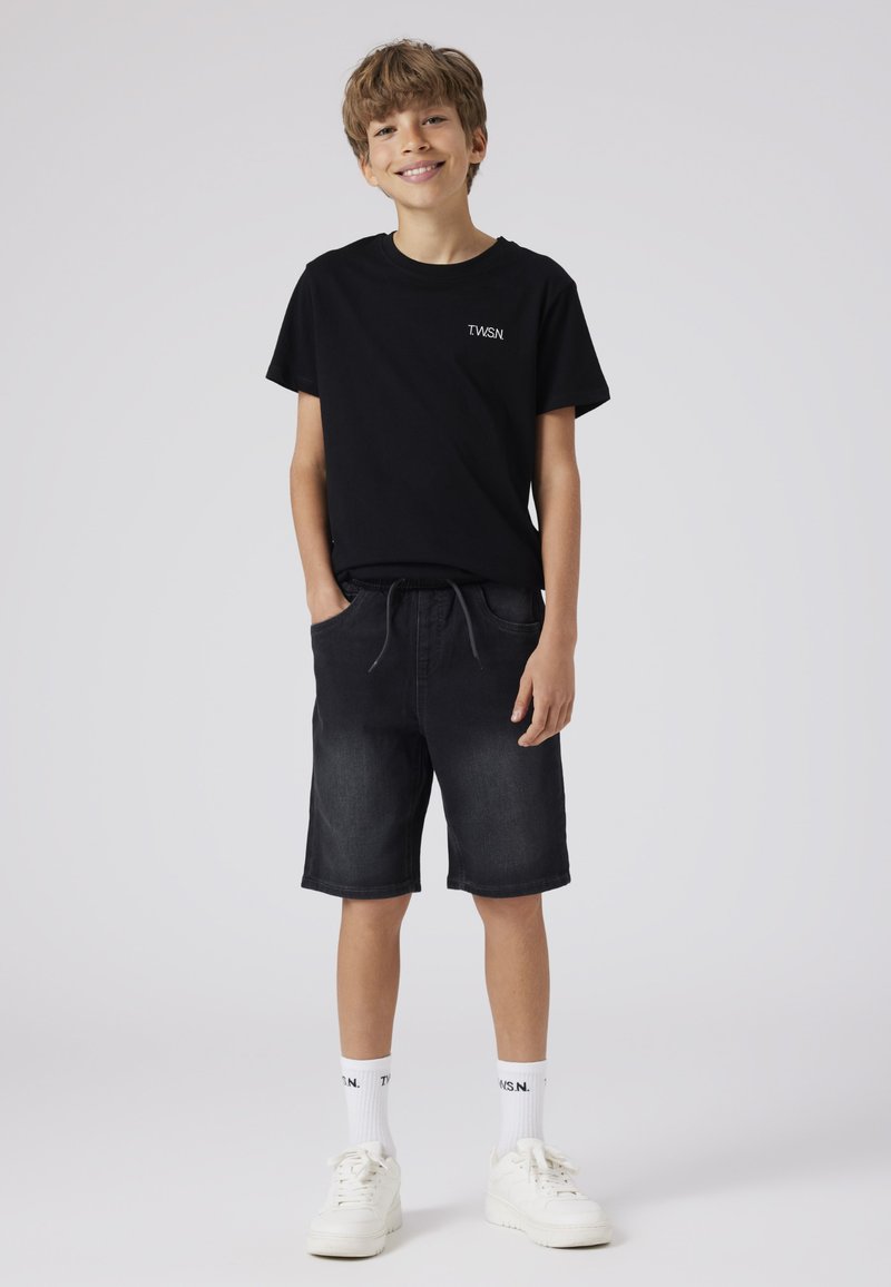 Juoda marškinėlių su logotipu, tamsūs džinsiniai šortai su raišteliu, balti sportbačiai ir baltos kojinės su juodu tekstu, pateikti ant paprasto fono.